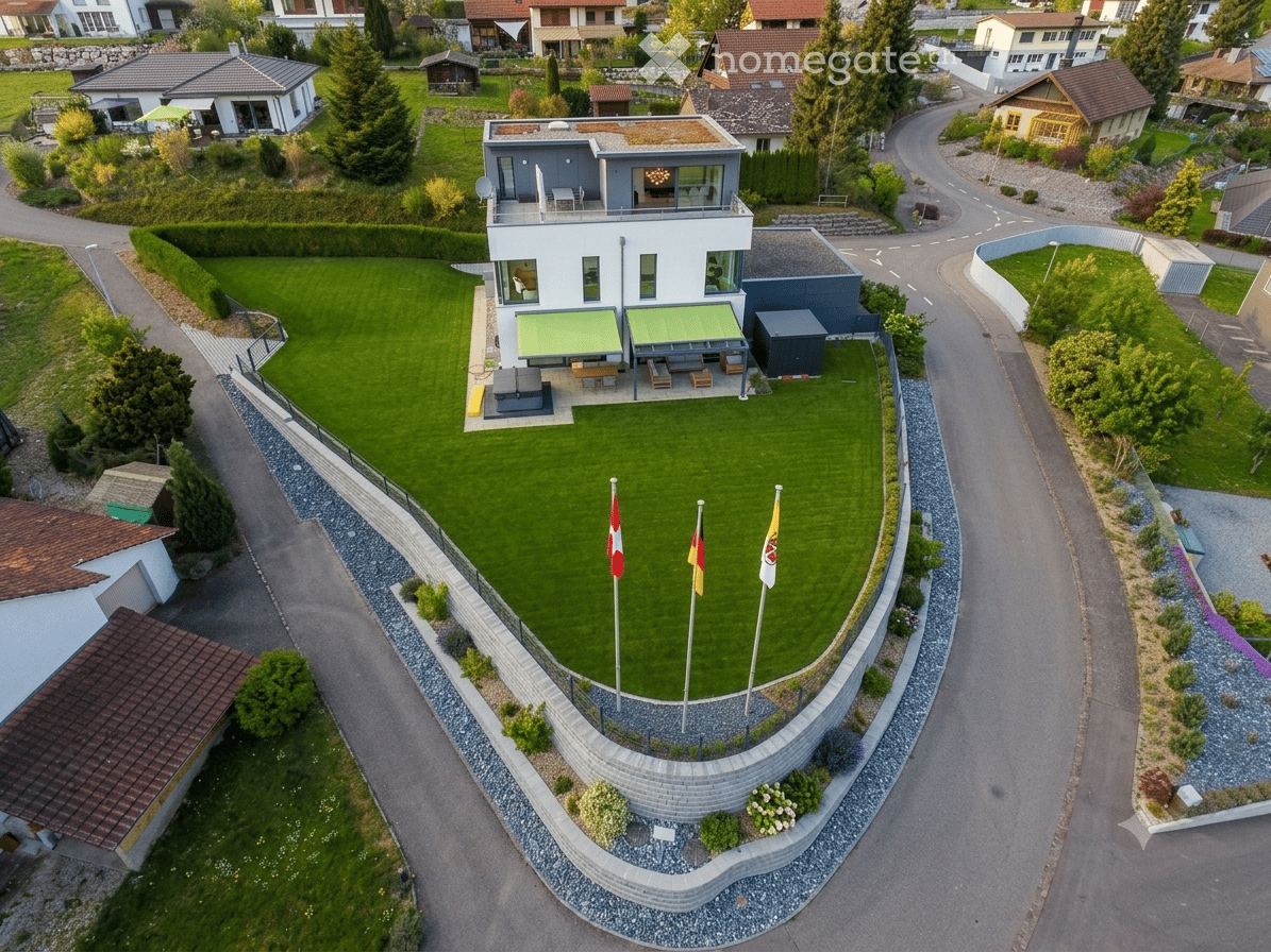 Aerial view of a modern house with a large, well-maintained lawn, surrounded by a stone wall and flags, with neighboring houses and a winding road visible.