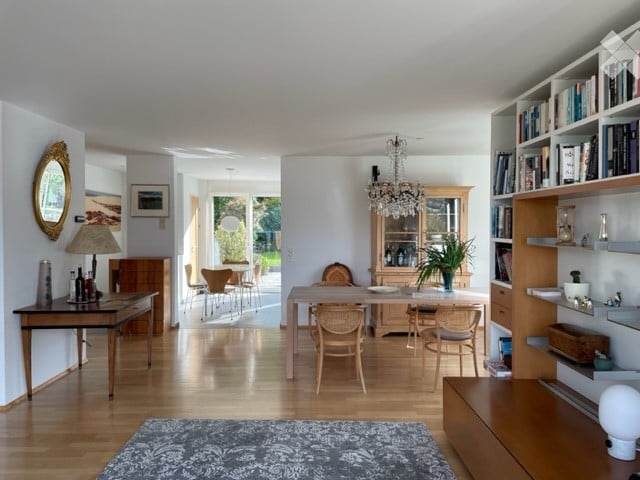 Bright, open-plan living and dining area with wooden flooring, large windows, and natural light. Features include a dining table with chairs, a bookshelf, a mirror, a lamp, and a cabinet with glassware.
