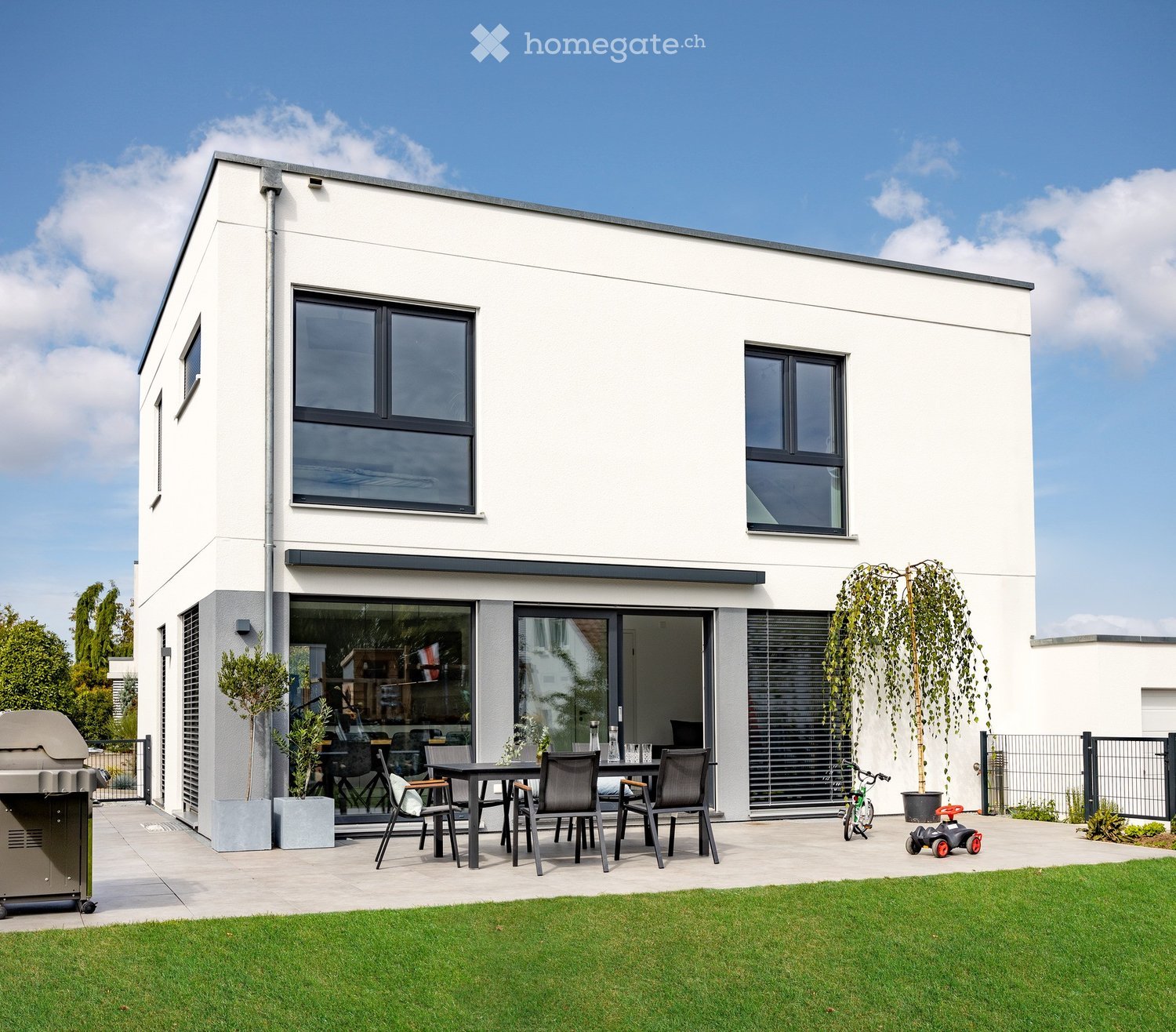 Modern house exterior with a spacious patio area featuring a dining set, grill, bicycle, and small toy car. The house has large windows and a clean, contemporary design.