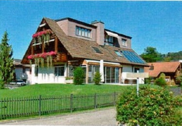 Two-story house with a pitched roof, solar panels, and a balcony with flower boxes. The house is surrounded by a well-maintained lawn and a metal fence.