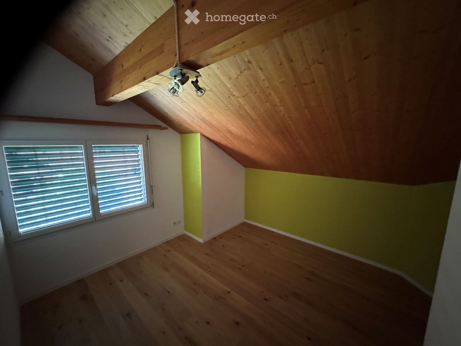 Empty attic room with wooden floors, exposed wooden beams, and a slanted ceiling. Two windows with blinds and two ceiling spotlights are visible.