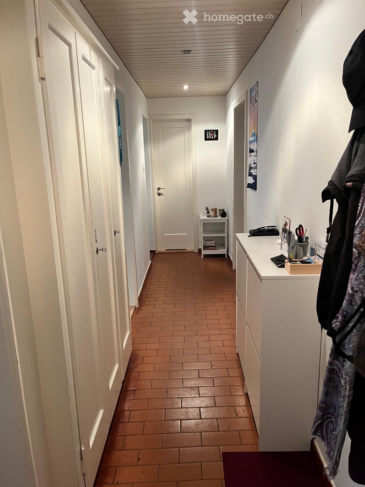 A narrow hallway with tiled flooring, white walls, and several doors. There is a small shelf with books and decorative items, and a coat rack with jackets and bags.