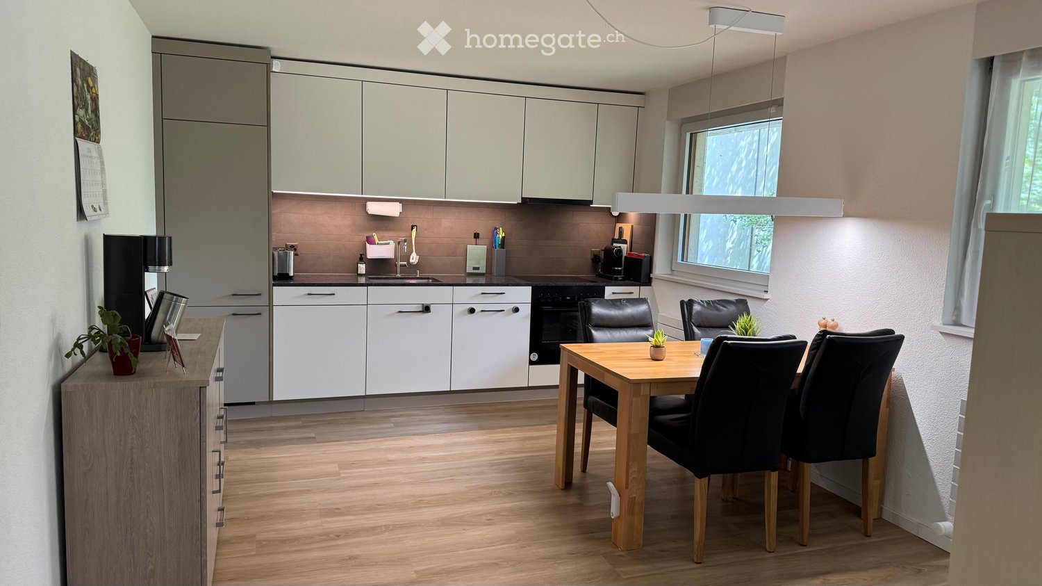 Modern kitchen with white cabinets, wooden dining table, four black chairs, and a small refrigerator. The space features a sleek design with a wooden floor and large windows.