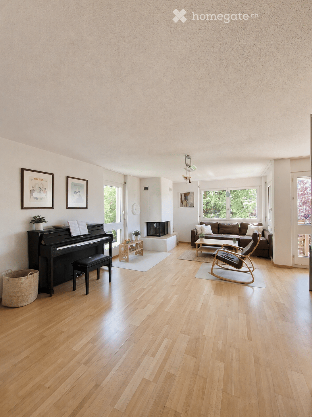 Spacious living room with wooden flooring, large windows, a brown sofa, coffee table, piano, fireplace, and decorative items.