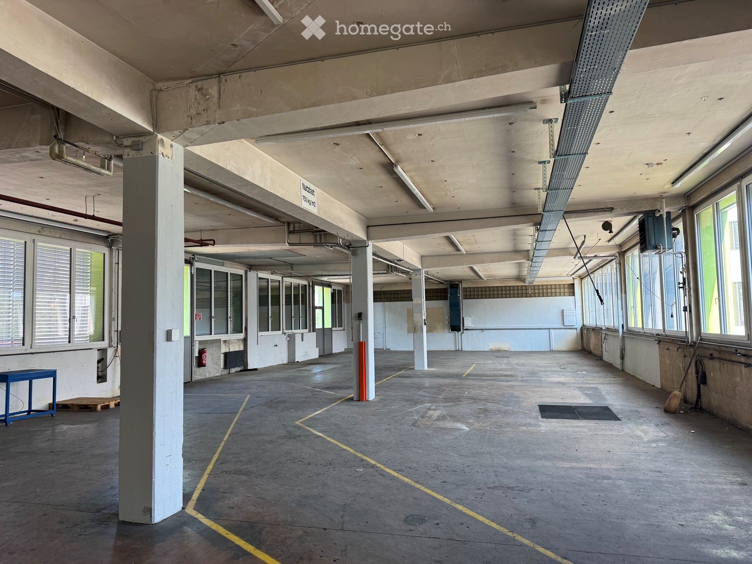 Empty industrial-style space with concrete floors, large windows, structural pillars, and minimal furnishings.