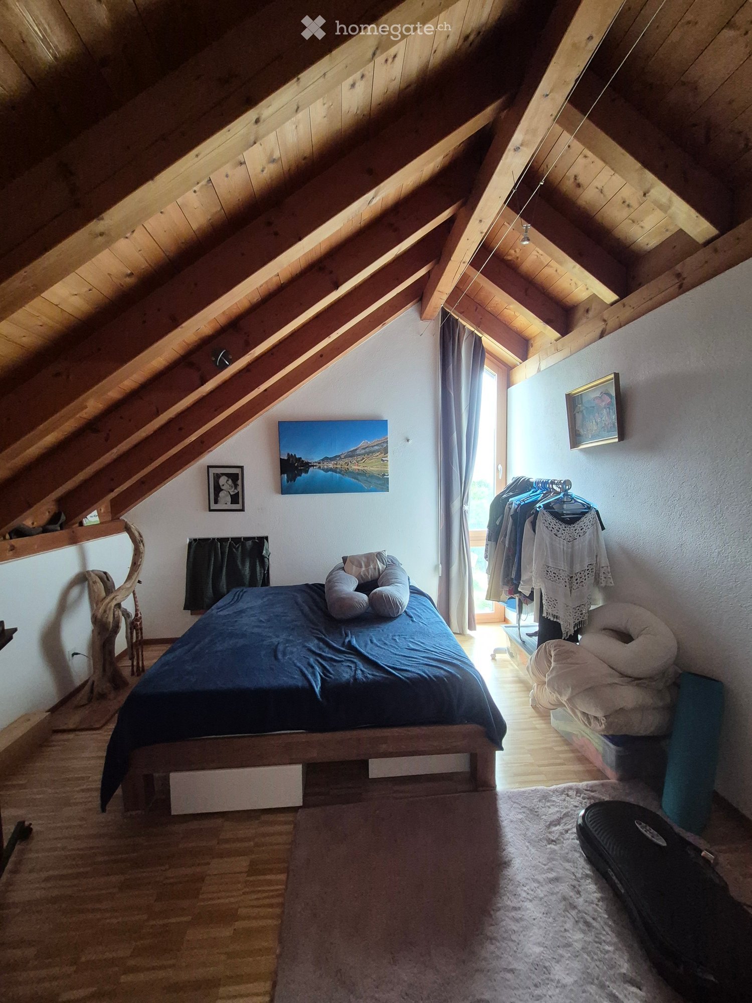 Cozy attic bedroom with wooden slanted ceilings, a large bed with blue bedding, pillows, a mirror, a clothing rack, and artwork on the walls.