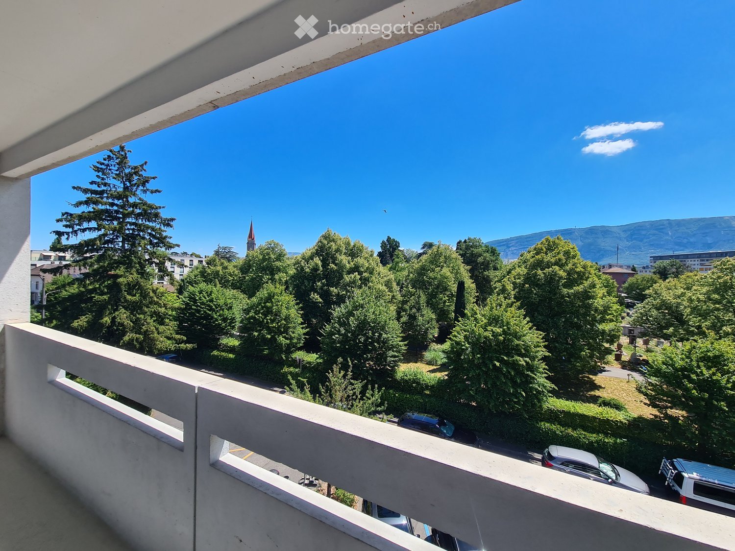 Balcony with a view of a park, trees, and distant mountains under a clear blue sky.