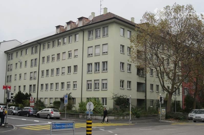 5-story building, many windows, some balconies, beige facade, greenery around, cars parked on street