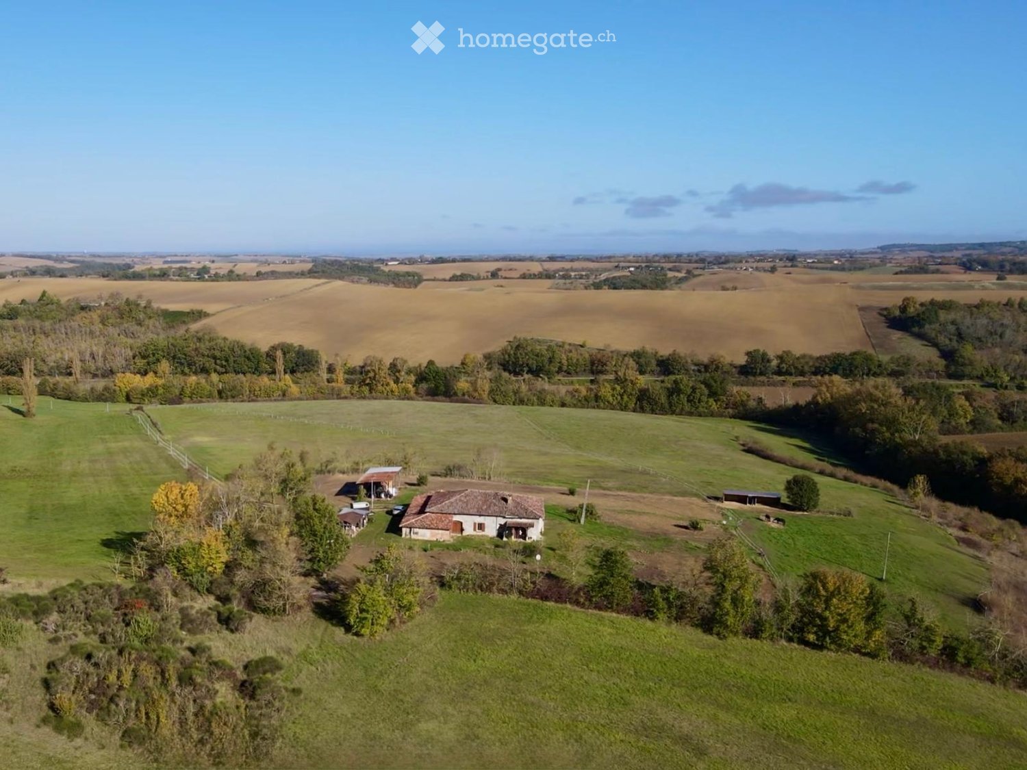 Aerial view of a rural house surrounded by green fields and trees, with a small outbuilding and a fenced area nearby.
