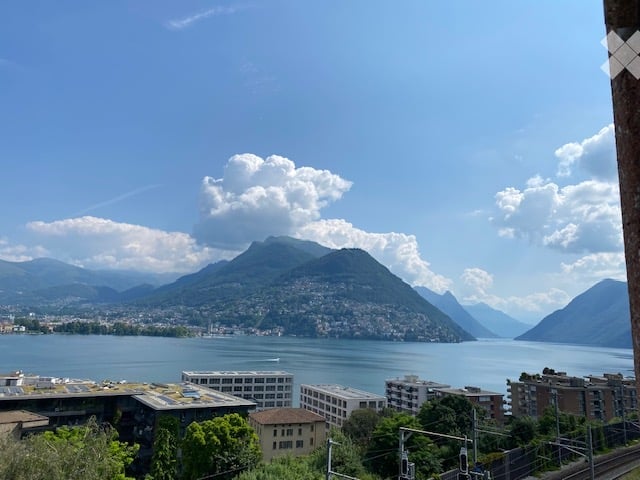 Panoramic view from a balcony showing a lake, mountains, and cityscape in the background.