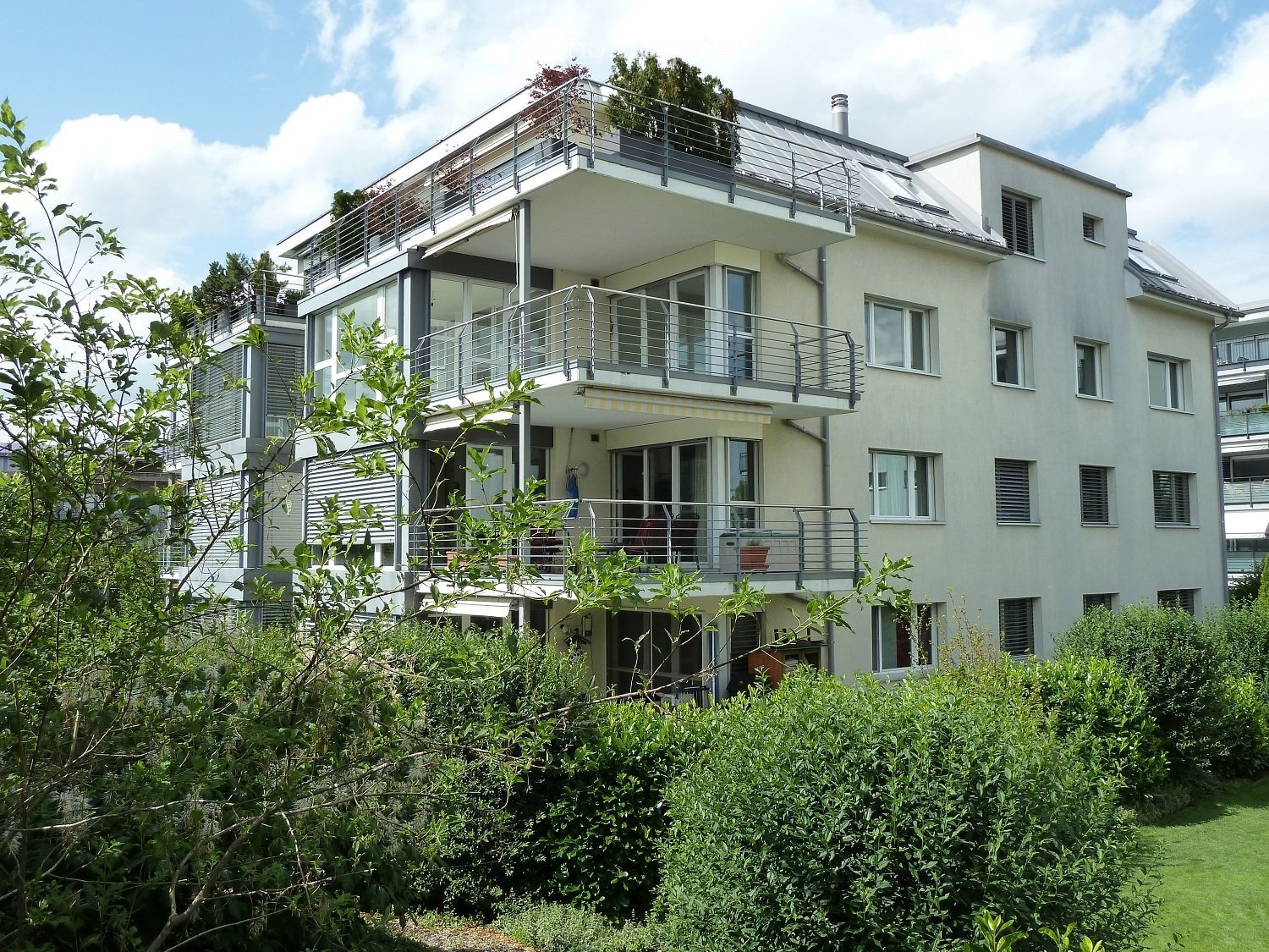 Modern multi-story residential building with balconies, large windows, and a well-maintained garden in front.