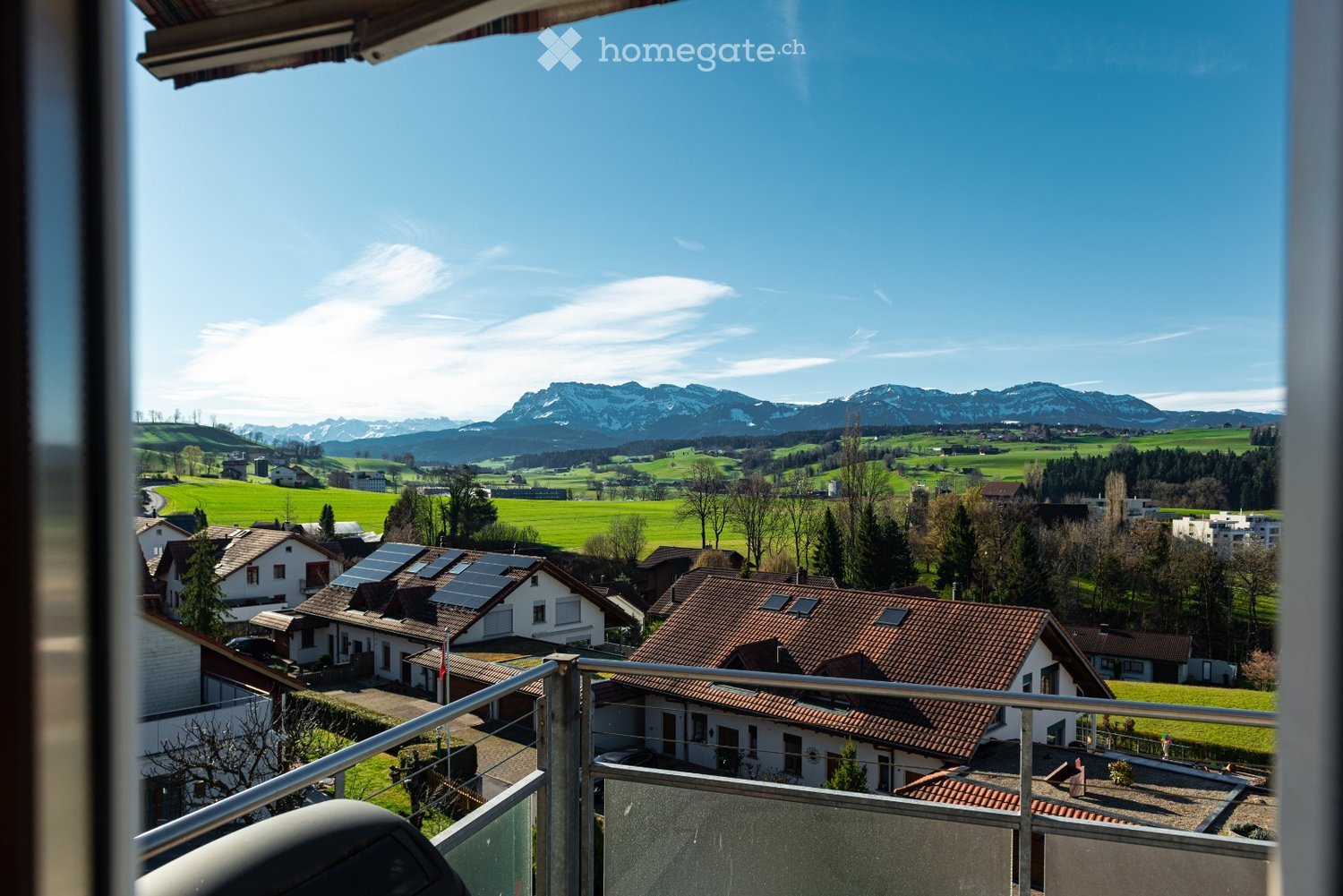 Balcony overlooking a scenic countryside with houses, green fields, and distant mountains under a clear blue sky.
