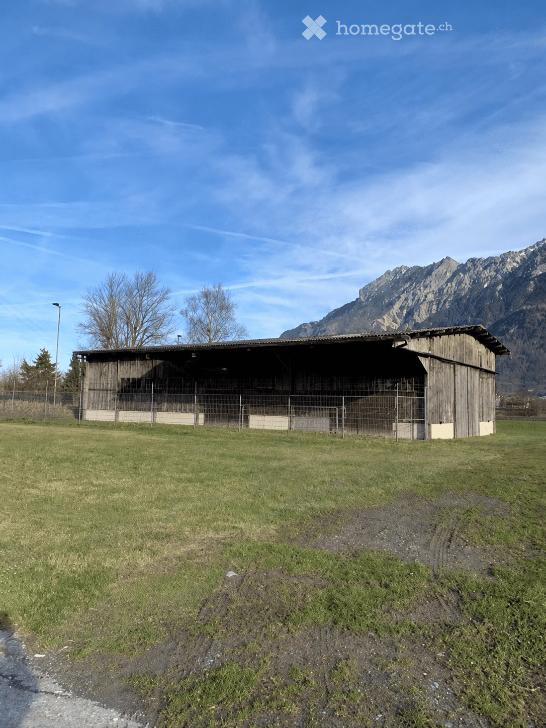 A wooden stable with a fenced area, situated in a grassy field with mountains in the background.