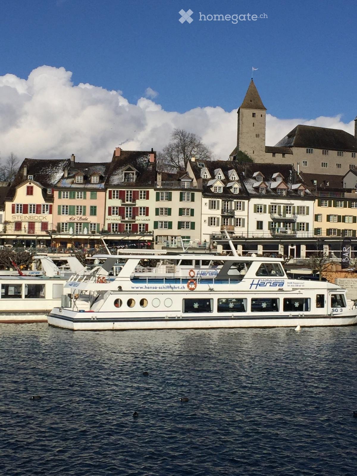 A scenic view of a town with traditional European architecture, featuring colorful buildings with red shutters and white facades, a prominent tower, and a boat on the water.