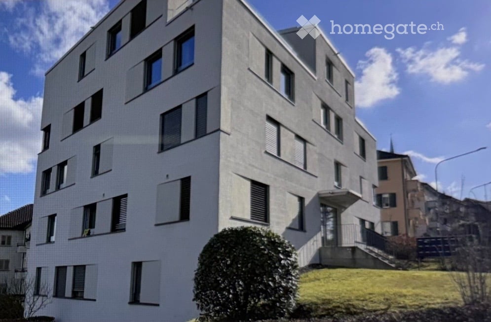 Modern multi-story residential building with white facade, numerous windows, and a small landscaped area in front.