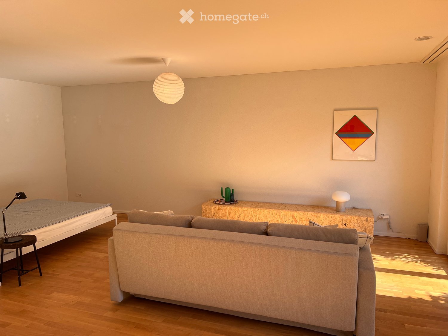 Modern living room with a sofa, bed, small table, and decorative items. Light wooden flooring and neutral-colored walls.