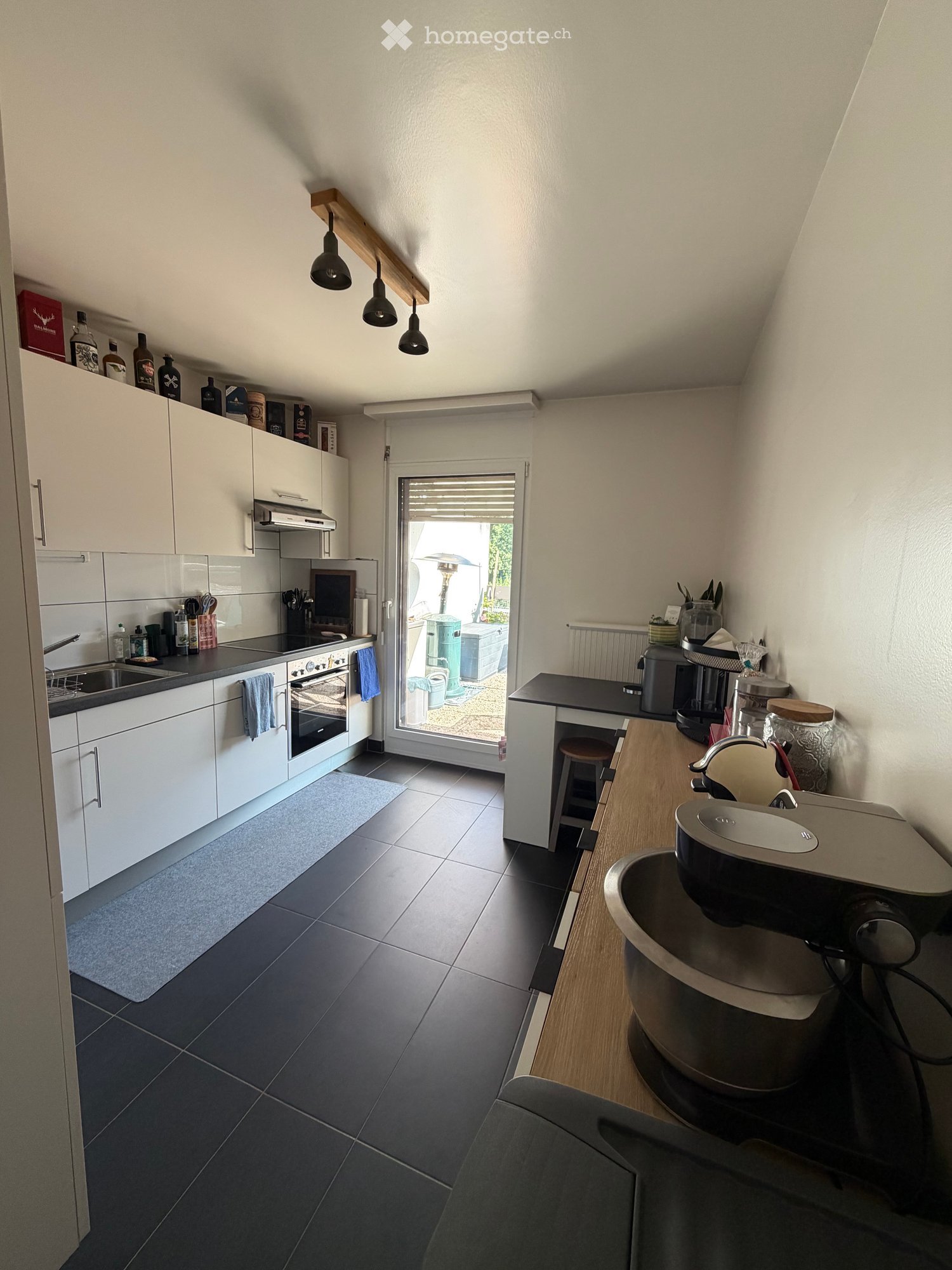 Modern kitchen with white cabinets, dark tiled floor, and a breakfast bar with a baby chair. Features include a sink, oven, and a coffee maker on the counter.