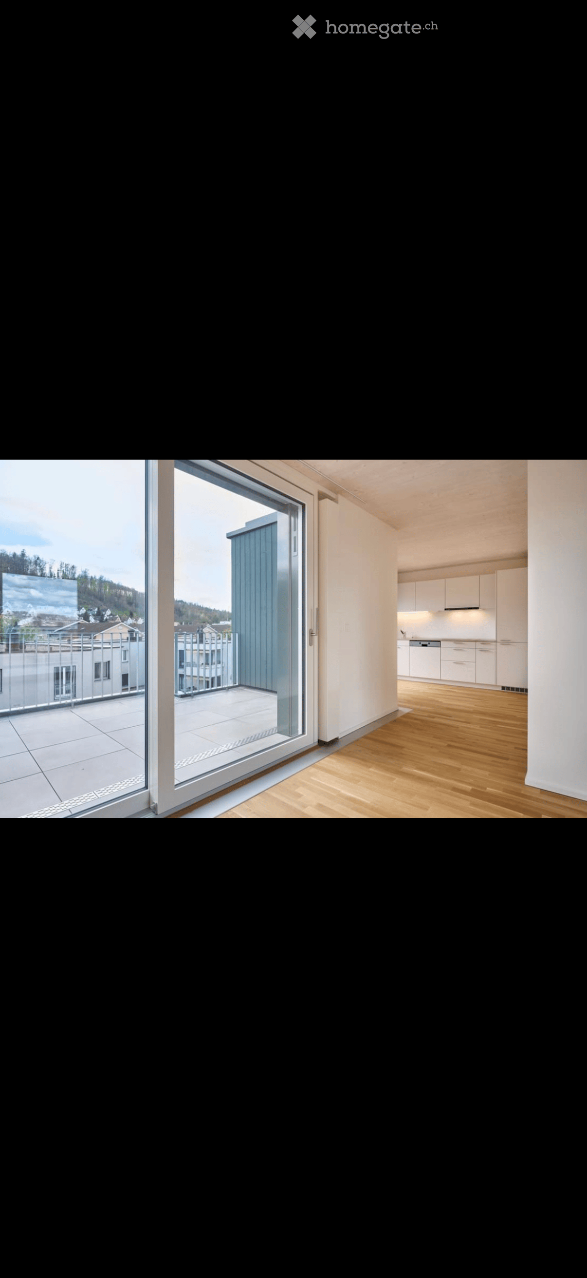 Modern interior with wooden flooring, large sliding glass door leading to a balcony, and a small kitchen area with white cabinets.