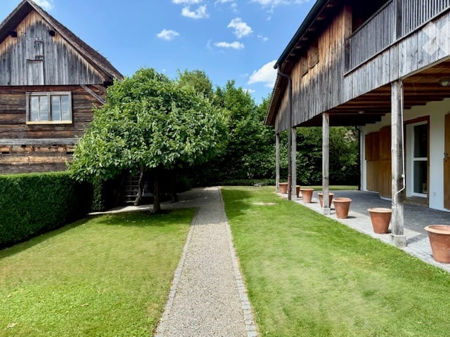 A well-maintained garden area with a gravel pathway, green lawn, and several wooden structures including a barn and a covered porch. Potted plants are placed along the pathway, and a large tree provides shade.