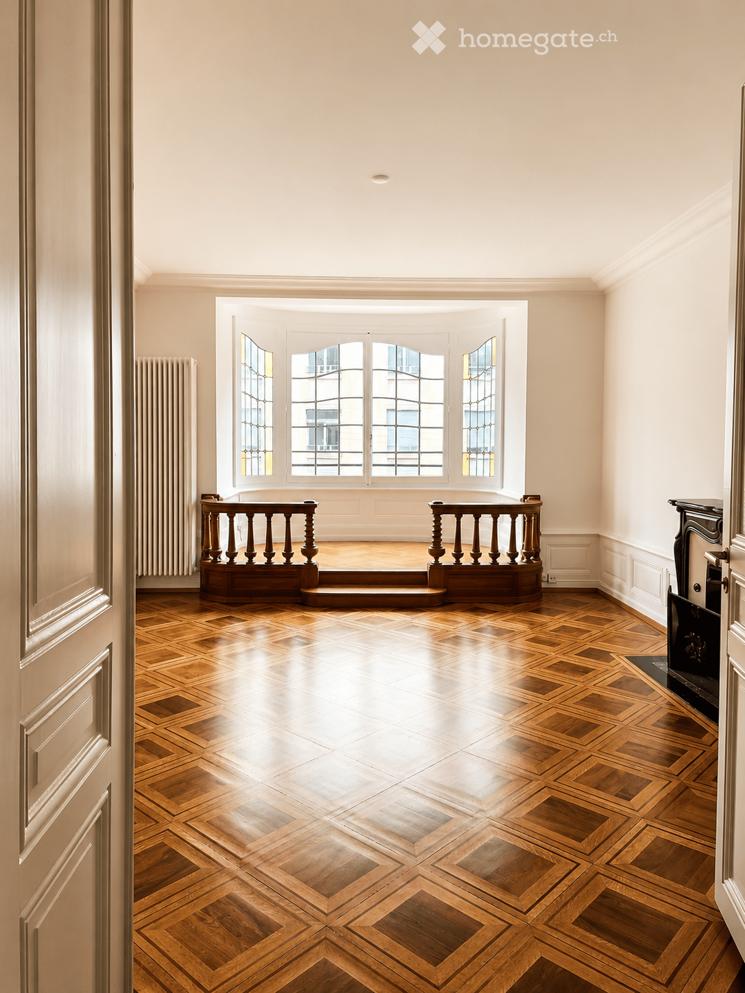 An empty room with wooden herringbone flooring, large windows with decorative glass, a raised platform with wooden balustrade, and a fireplace on the right side.