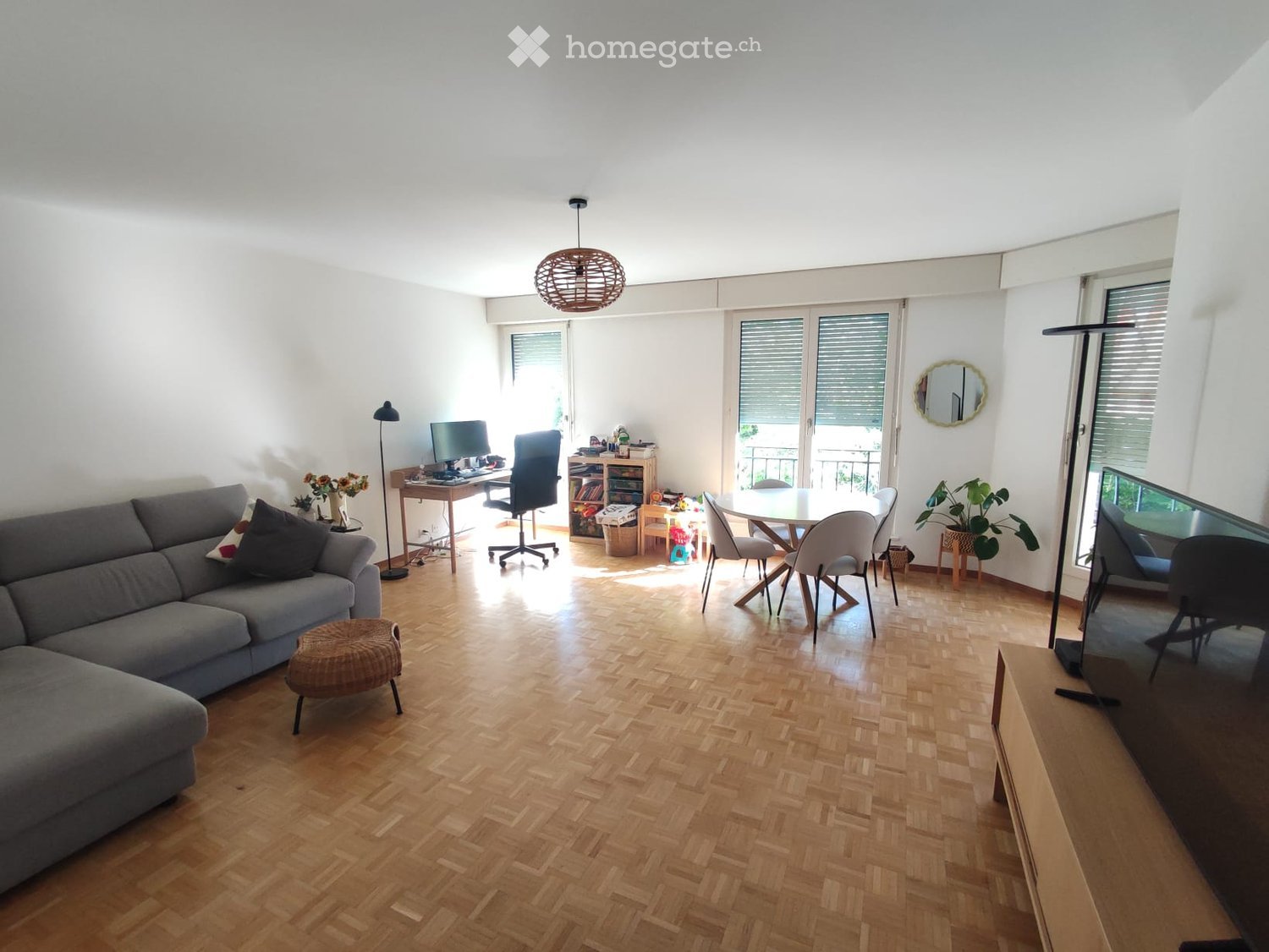 Bright living room with wooden flooring, large windows, and a balcony. Features a grey sofa, a round dining table with chairs, a TV screen, and a desk with a computer.