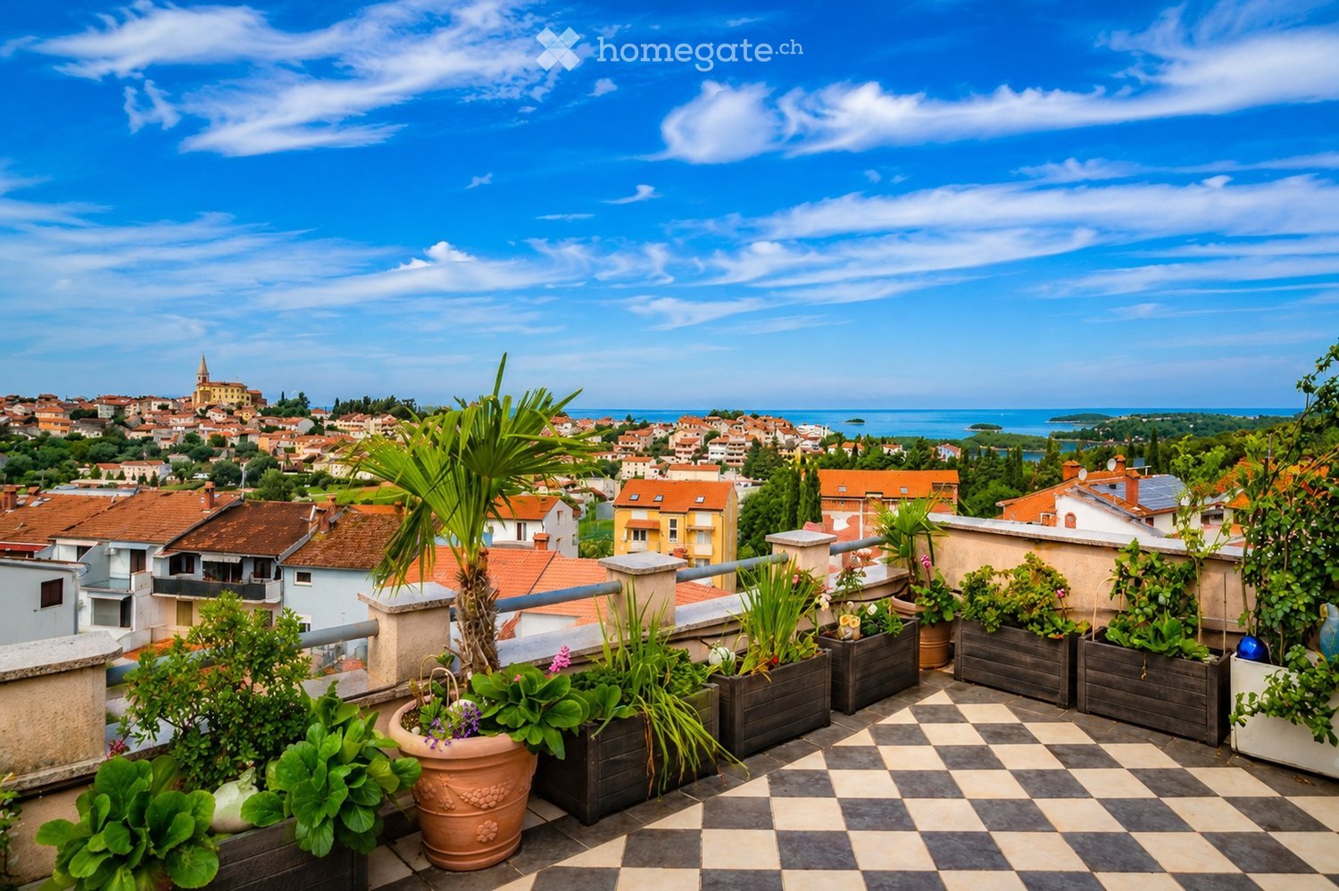 A rooftop terrace with potted plants and a checkered tile floor, overlooking a town with red-tiled roofs and a distant sea view.