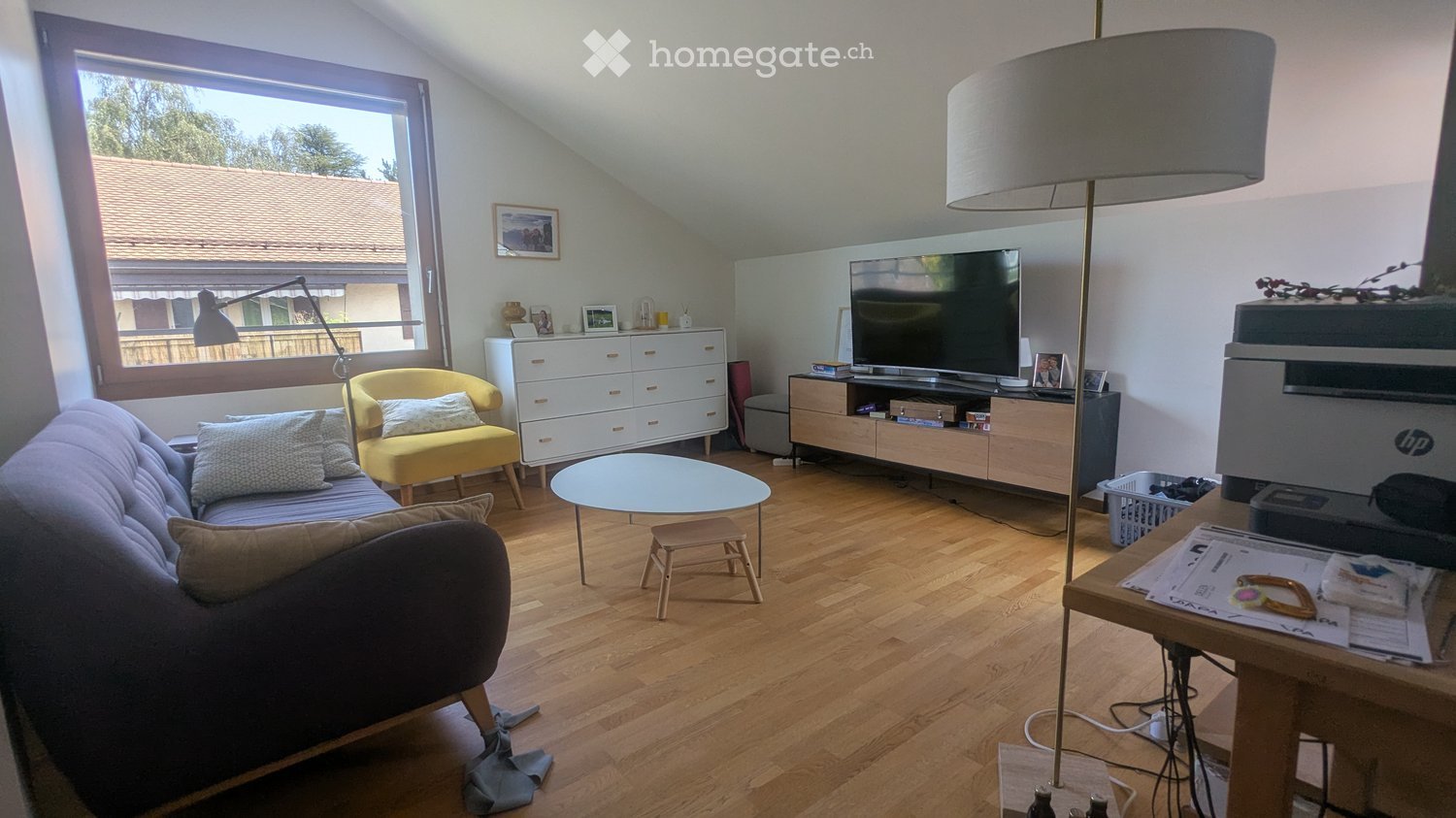 Living room with wooden flooring, large window, sofa, armchair, coffee table, TV, floor lamp, and printer on a table.