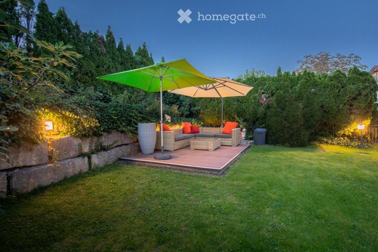 Well-maintained garden with a raised wooden deck, outdoor sofa set, coffee table, large umbrella, and decorative vase.