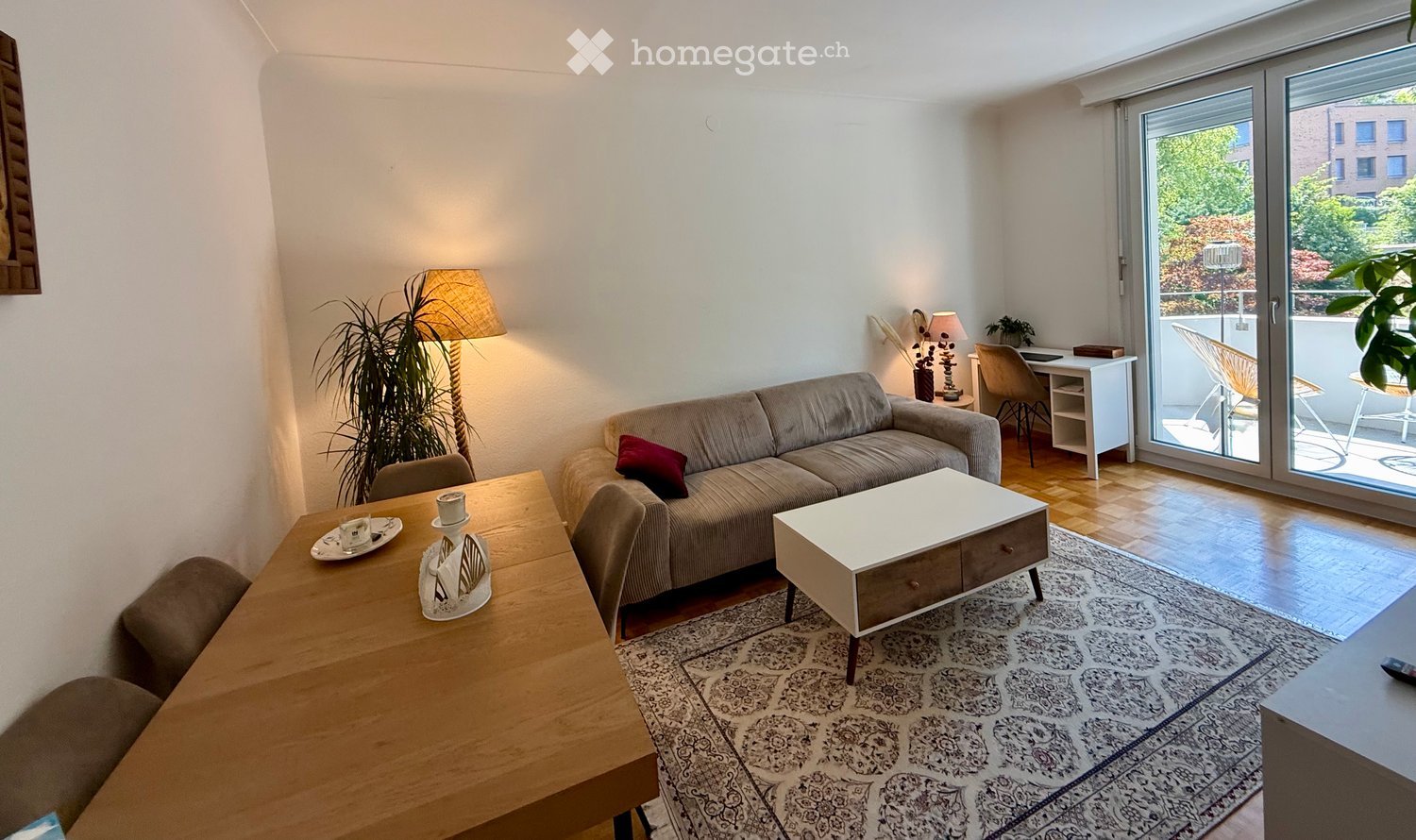 Bright living room with a grey sofa, coffee table, dining table, chairs, floor lamp, desk, and plants. Large sliding glass doors lead to a balcony.