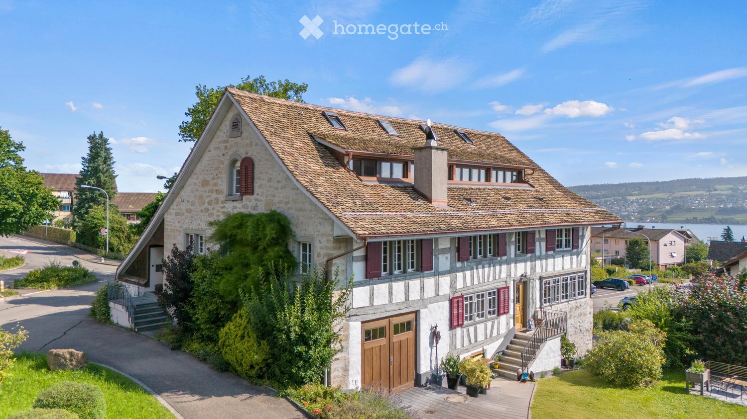 A two-story stone house with a tiled roof, multiple windows with shutters, and a wooden garage door. The house is surrounded by greenery and has a paved driveway leading to the entrance.