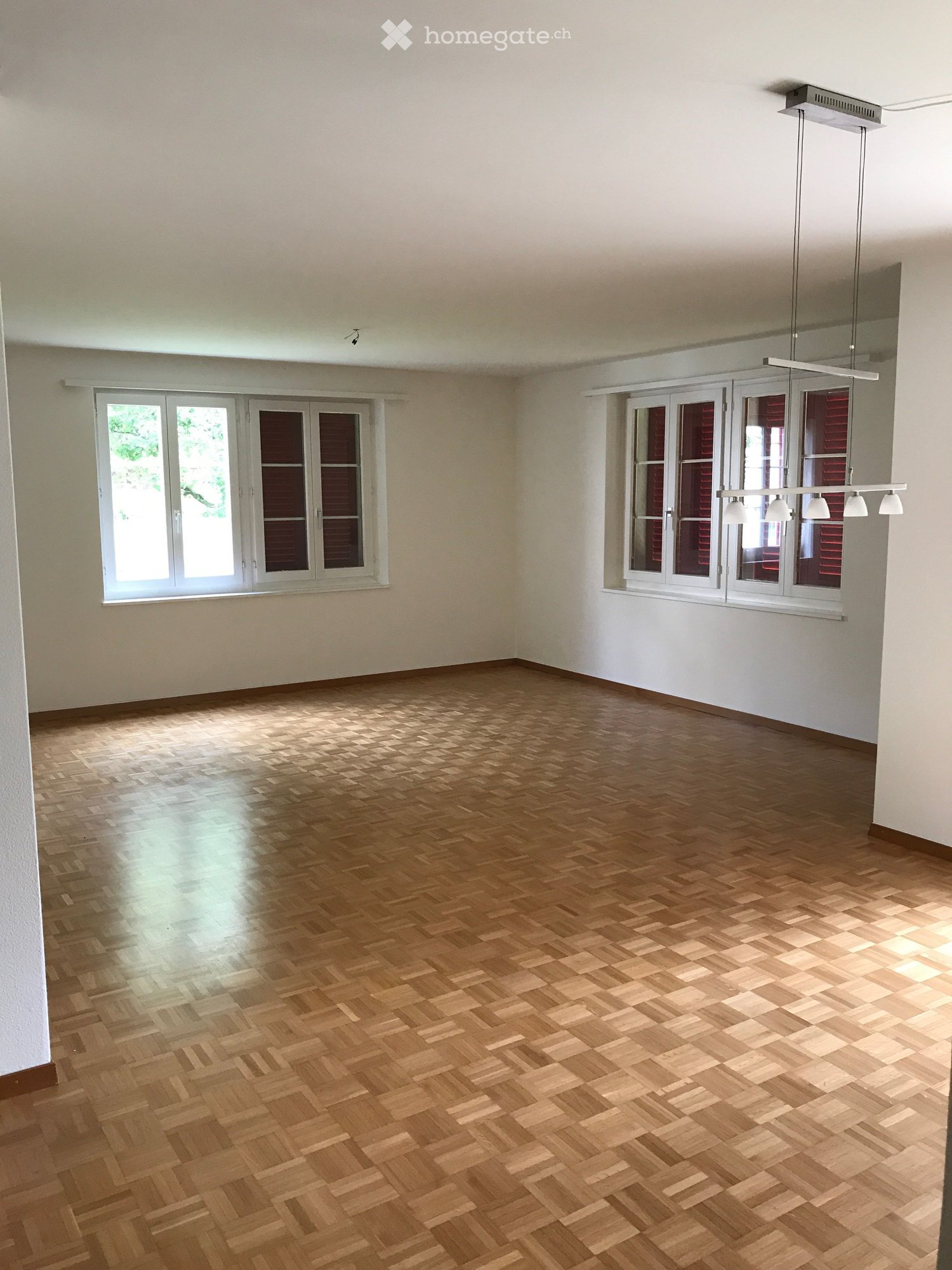 Empty room with wooden herringbone flooring, white walls, and large windows with white frames. Natural light streams in, illuminating the space.