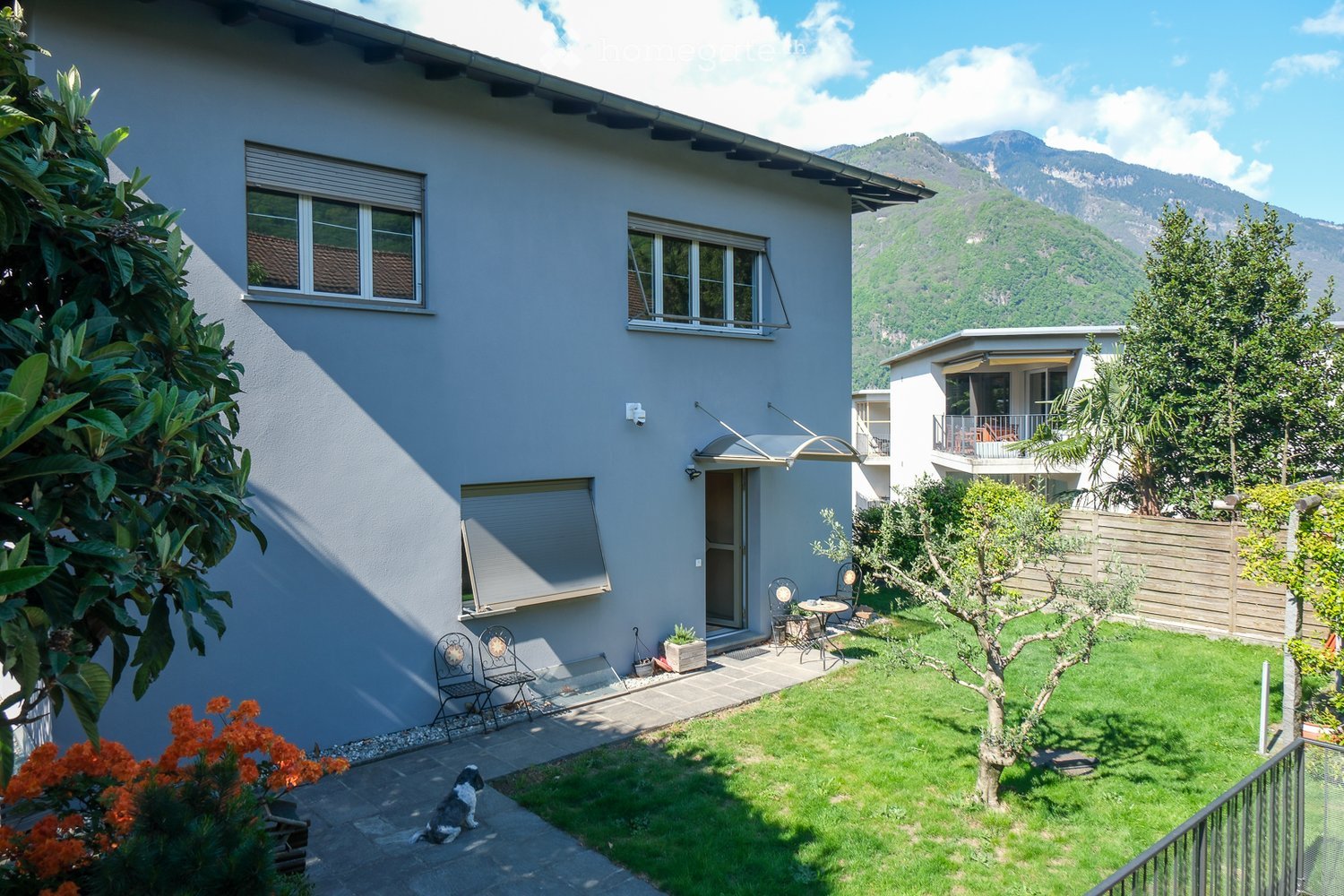 A well-maintained garden area with a small patio, chairs, a table, and a dog sitting on the ground. The garden is surrounded by a wooden fence and features a lawn, trees, and a view of mountains in the background.