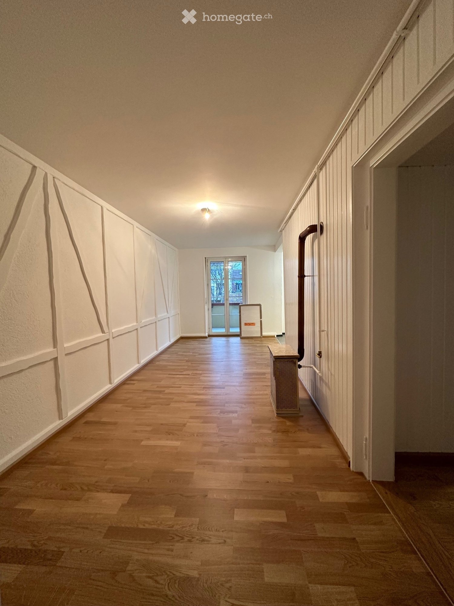 Empty hallway with wooden flooring, white walls, and a ceiling light. A door at the end leads to another room.