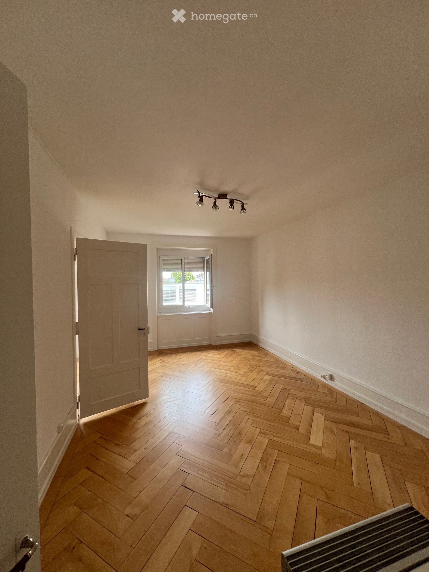 Empty room with wooden herringbone flooring, light-colored walls, a ceiling light fixture, a window with blinds, and a partially visible radiator.