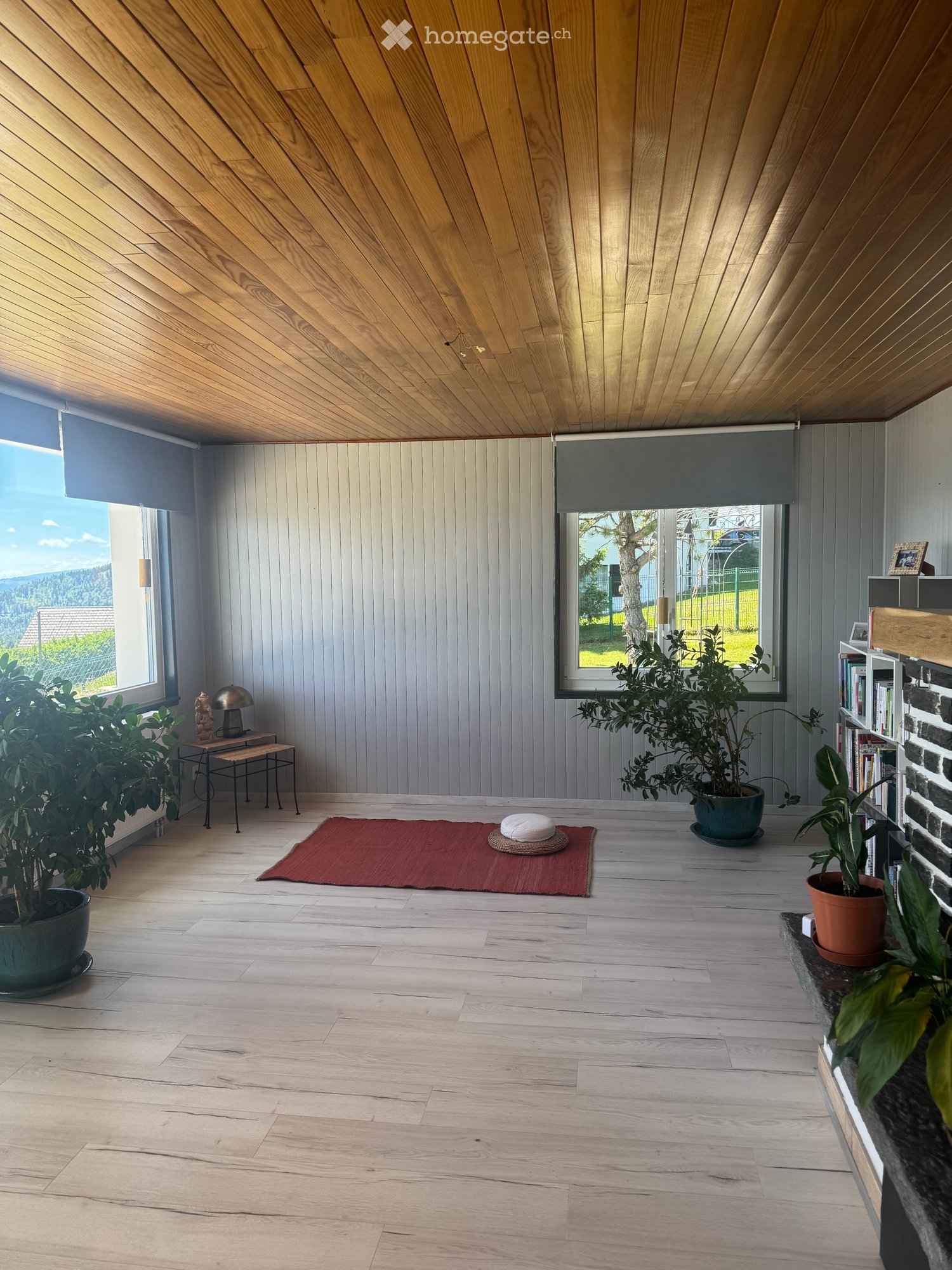 Bright, minimalist room with wooden floors and a slanted wooden ceiling. Features large windows with roller shades, a small table with a lamp, a bookshelf, potted plants, and a red meditation mat with cushions.