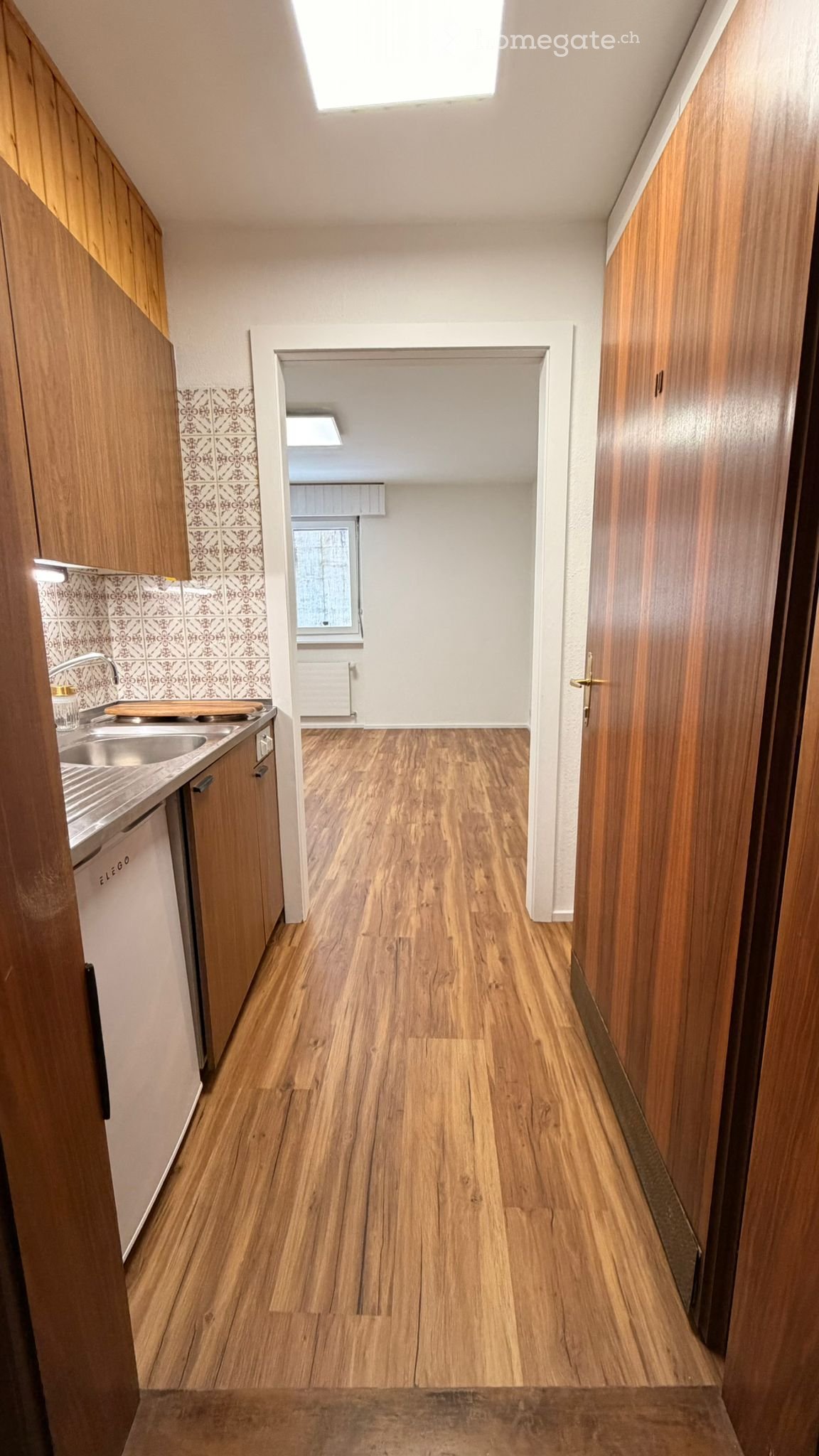 A narrow hallway with wooden flooring and walls, leading to a kitchen area with wooden cabinets, a sink, and a window.