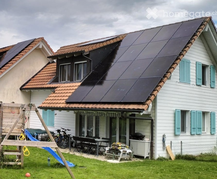 Detached house with solar panels, brown roof, white walls, wooden door, garden with slide, bicycles, table and chairs, grill
