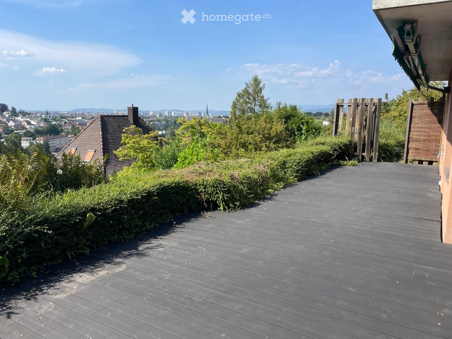 Wooden deck with a view of the city and surrounding greenery, featuring a wooden railing and a pathway.