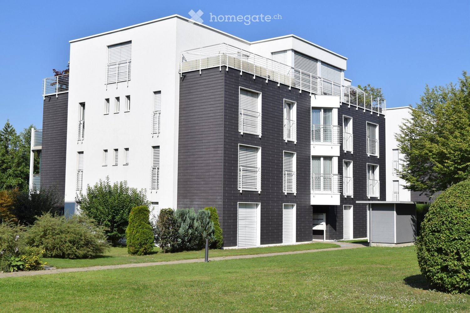 Modern two-story building with white and dark gray facade, multiple windows, balconies, and surrounding greenery.