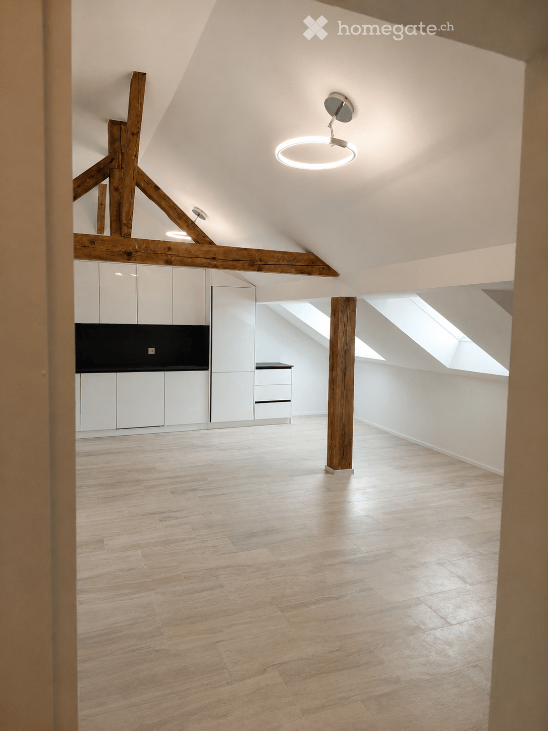 Bright, open attic space with wooden beams, light wooden flooring, white walls, and built-in white cabinets. Features skylights and a modern circular ceiling light fixture.