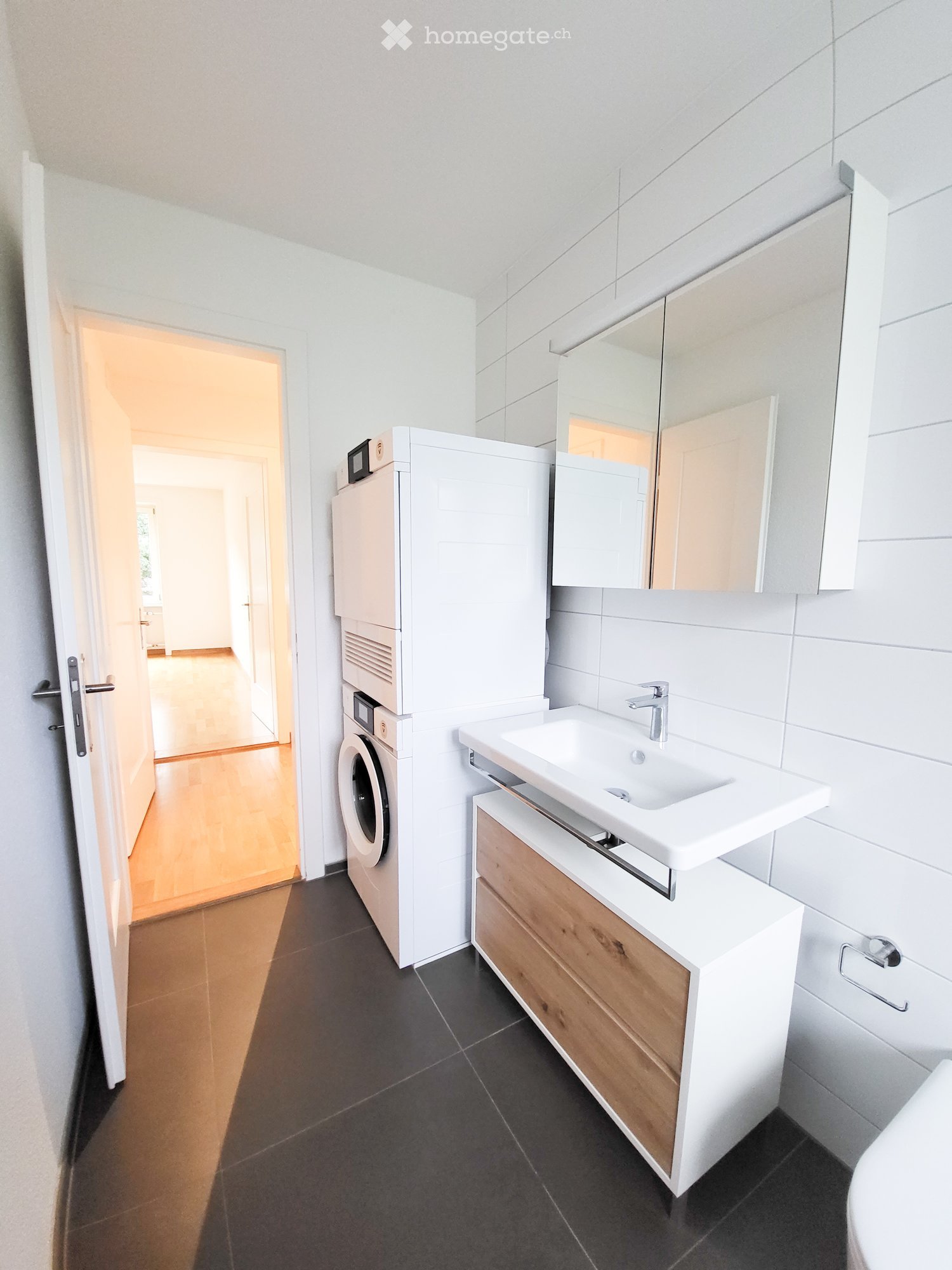 Modern bathroom with a white sink, wooden cabinet, mirror, and a washing machine. The floor is dark tiled, and the walls are white.
