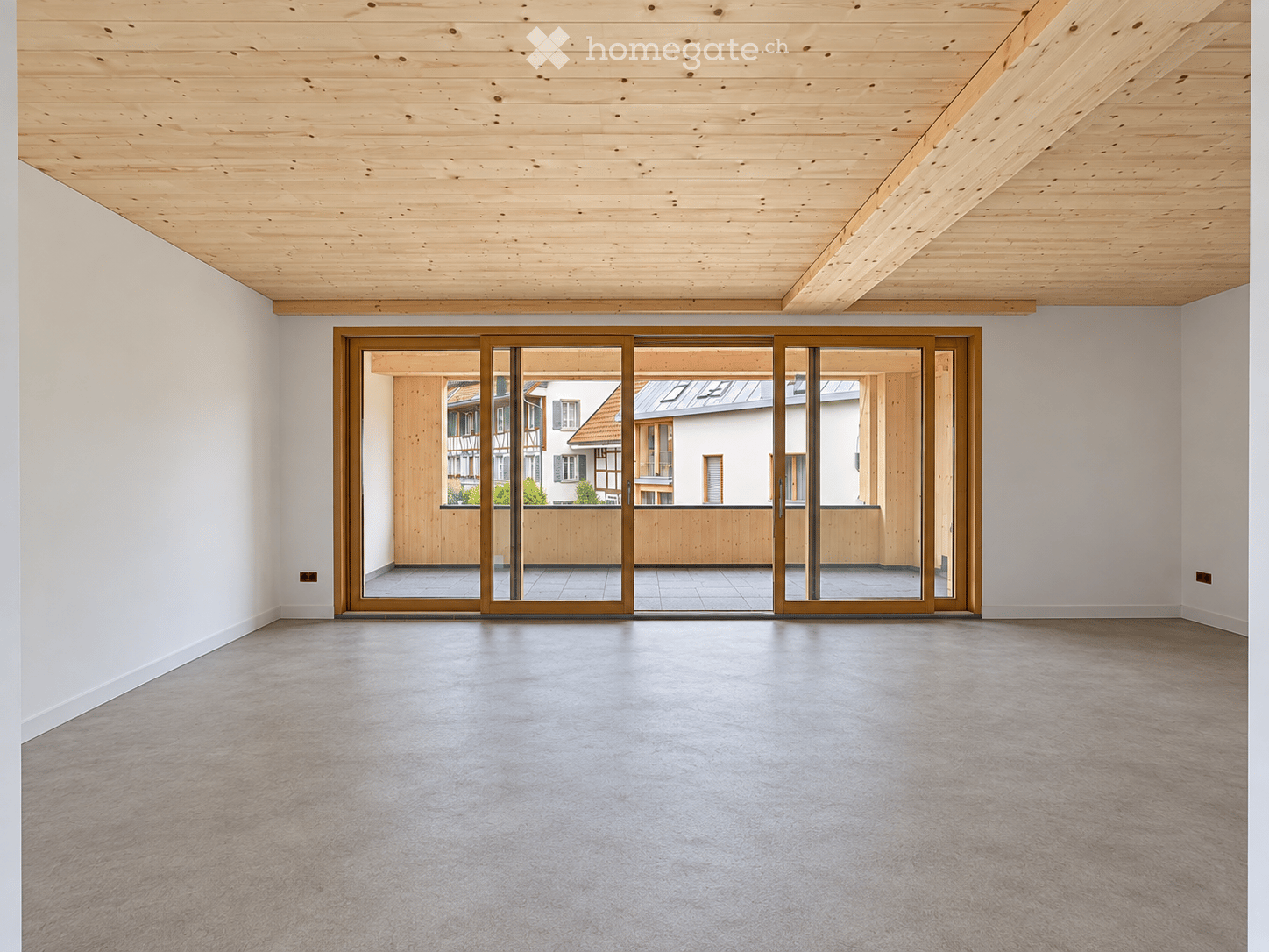 An empty room with a wooden ceiling, grey concrete floor, and large sliding glass doors leading to a wooden deck and outdoor area.