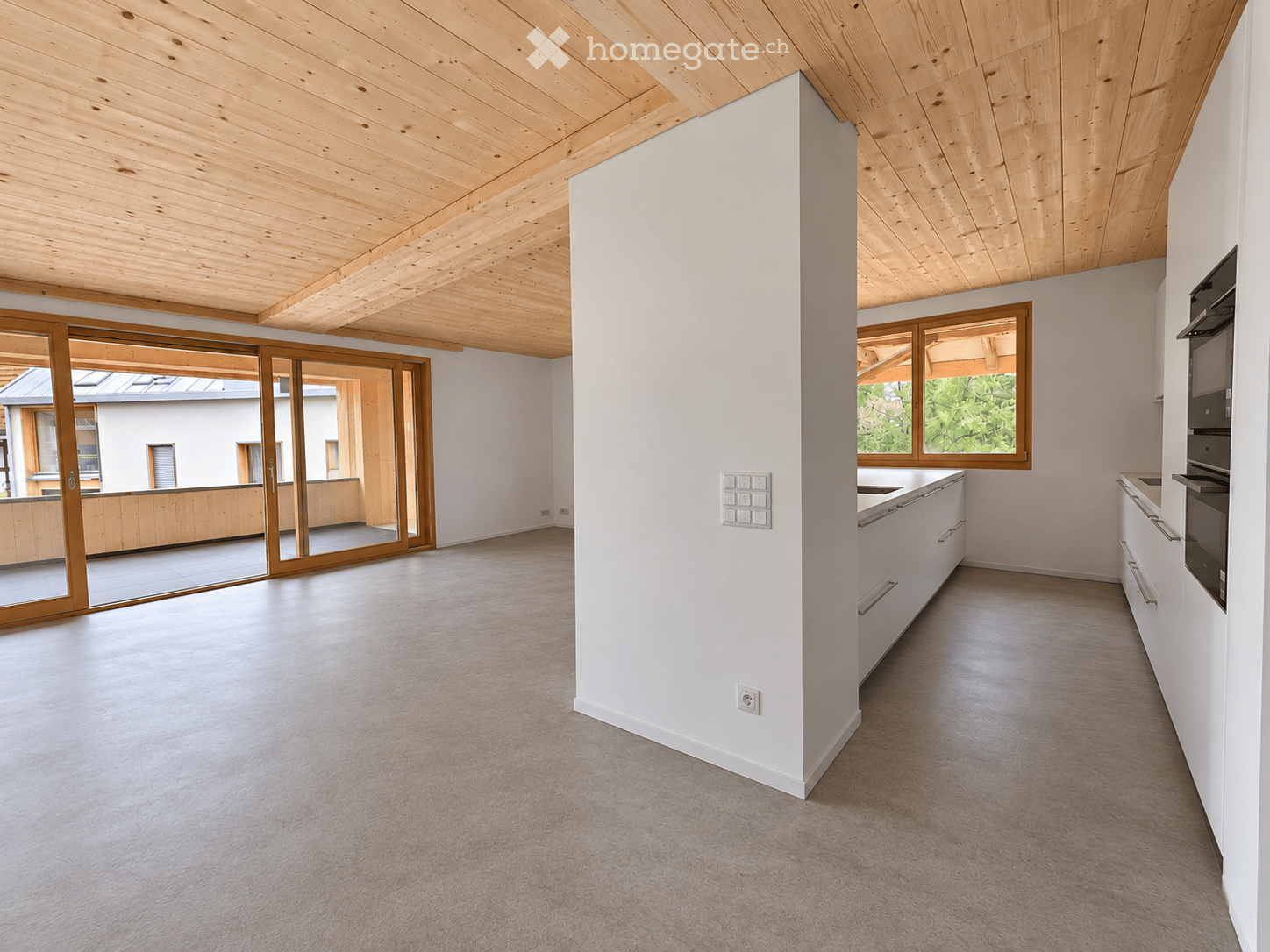Open-plan space with light-colored walls, wooden ceiling beams, and carpeted floor. Features a modern kitchen area with white cabinets and stainless steel appliances, large windows, and sliding glass doors leading to an outdoor area.