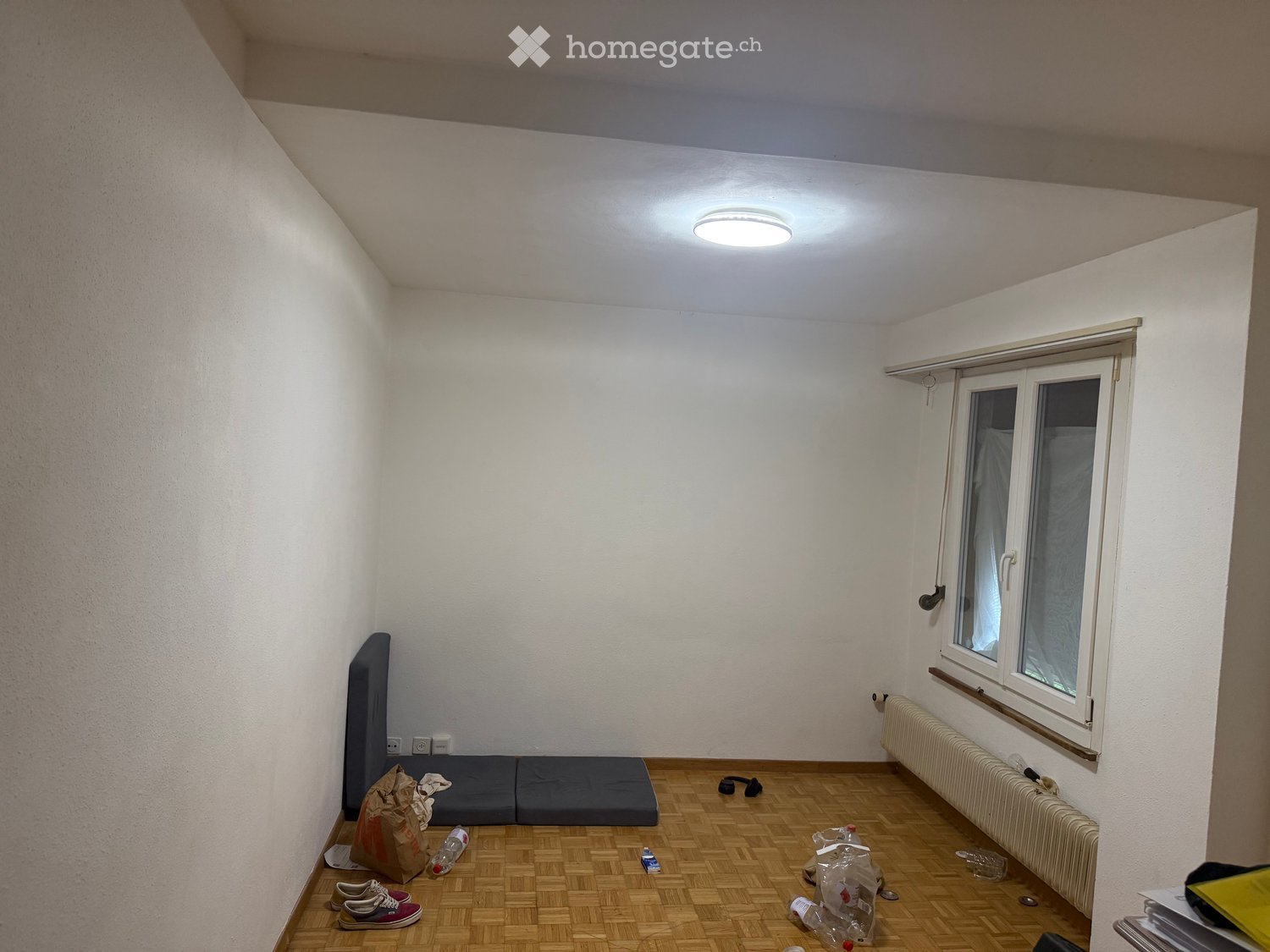 Empty room with wooden flooring, white walls, a window with blinds, and a radiator. No furniture or major items present.
