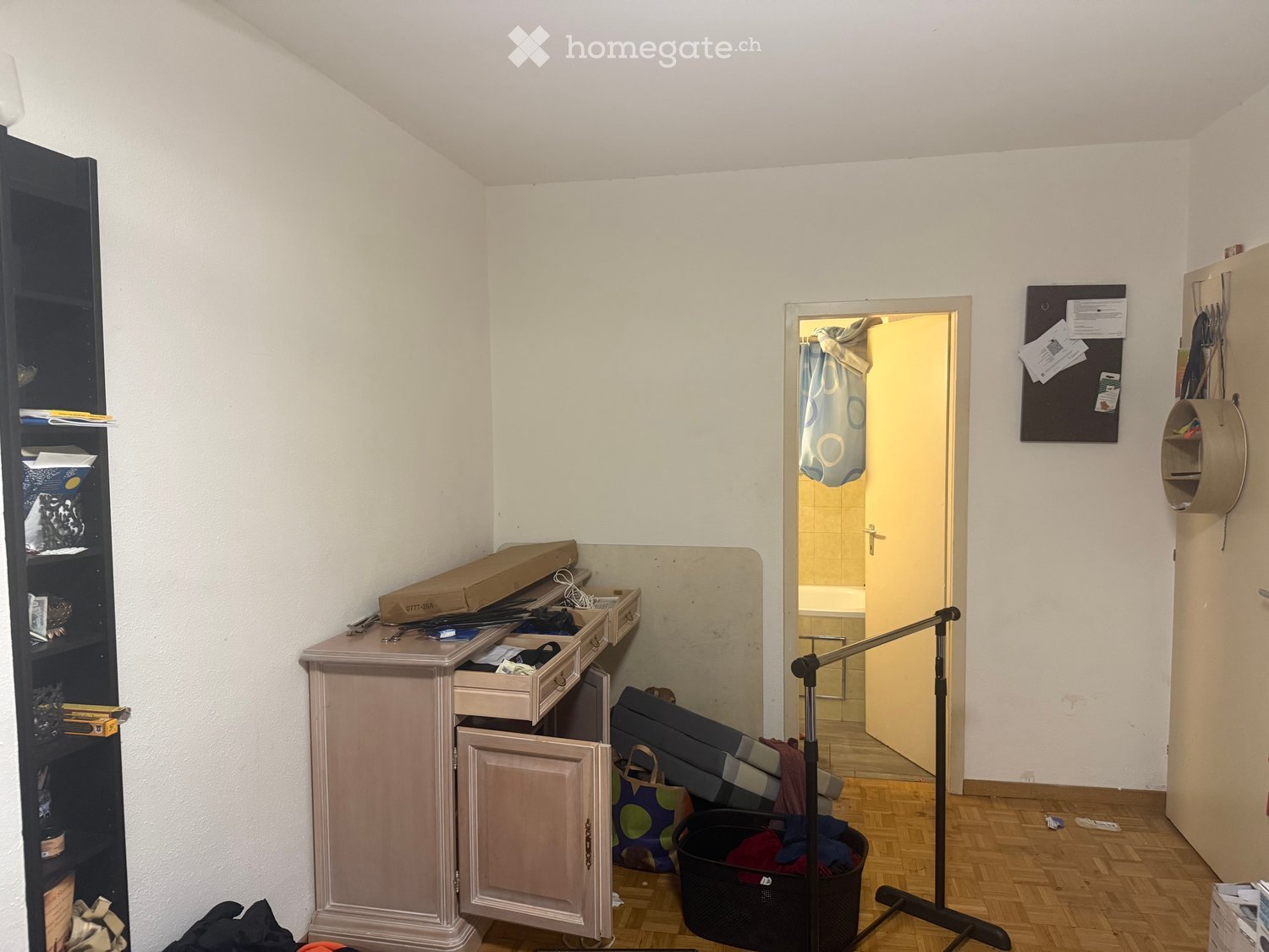 The room has a wooden floor, a cabinet with drawers, a shelf, a mirror, a basket, and a stool. There is an open door leading to a bathroom with a shower curtain visible.