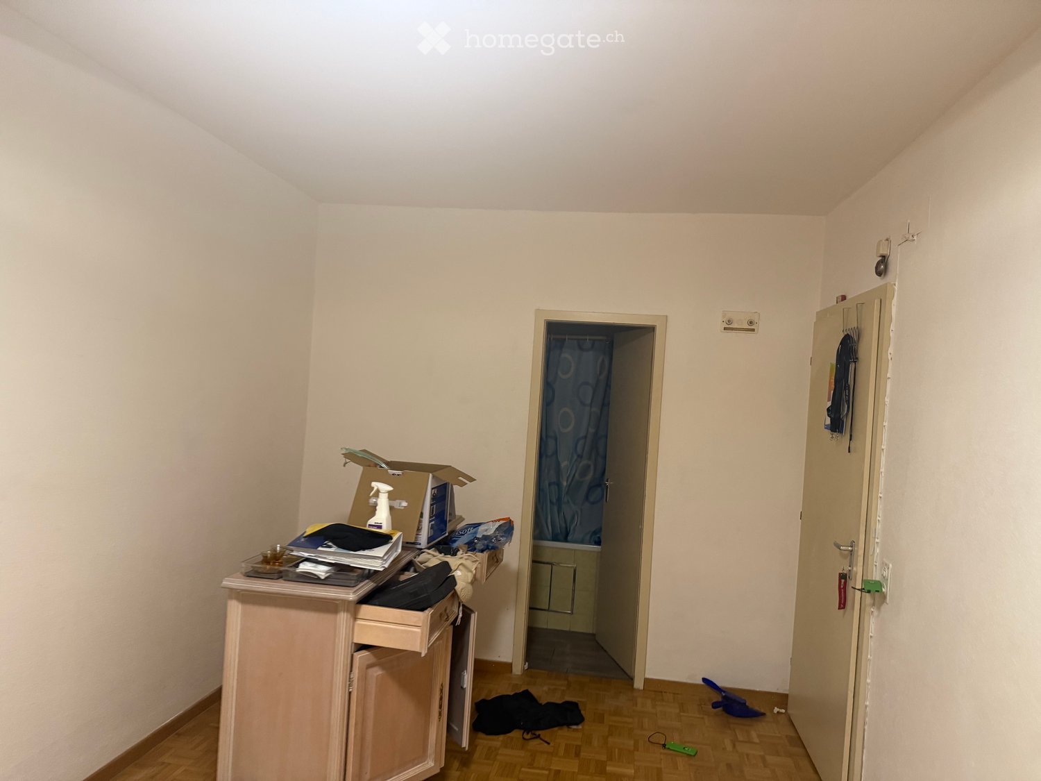 A small, unfurnished room with a wooden floor, a table with various items, a door leading to a bathroom, and a curtain visible in the background.