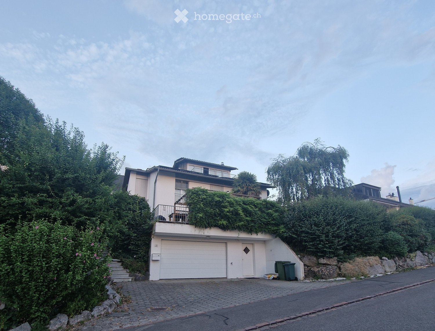 Two-story house with a garage, balcony, and lush greenery on the roof and surrounding area.