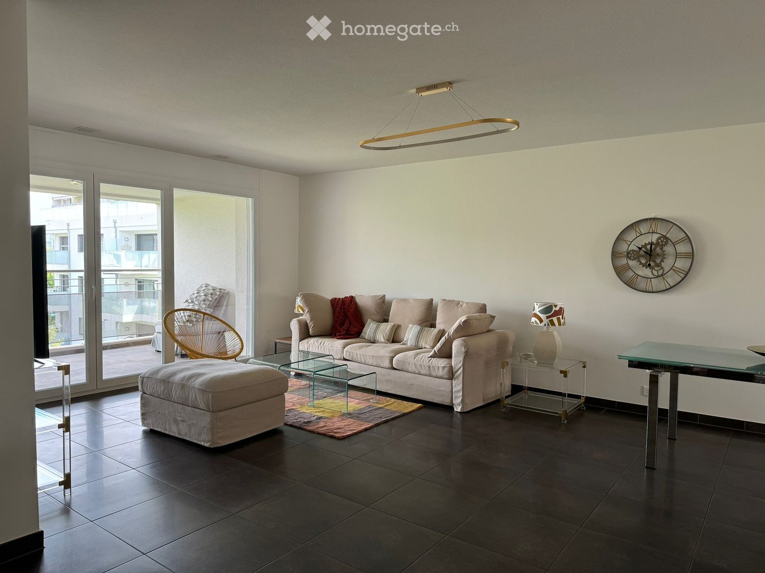 Modern living room with a beige sofa, ottoman, glass coffee tables, a wicker chair, a glass side table with a lamp, and a decorative wall clock. Large sliding glass doors lead to a balcony.