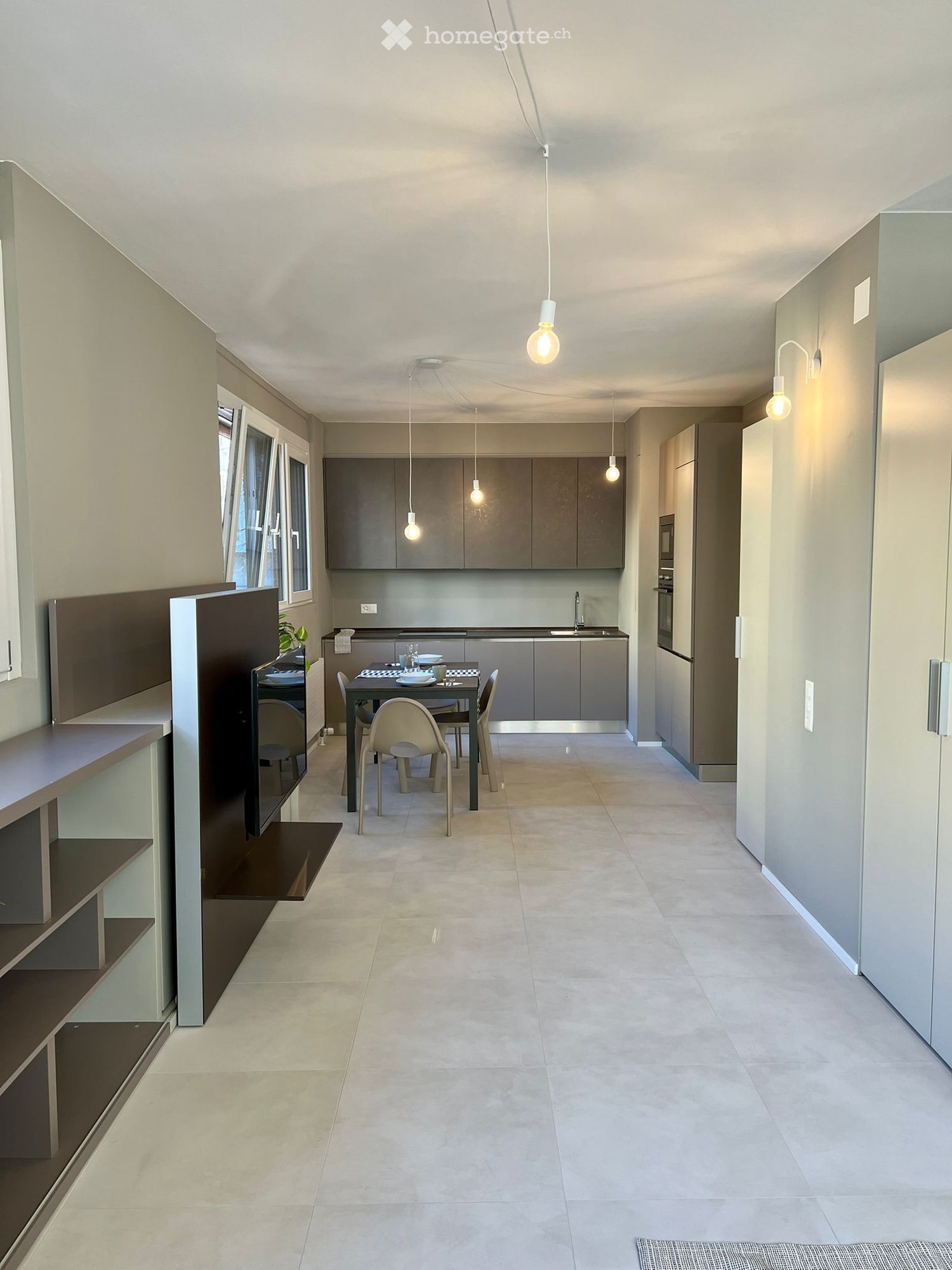 Modern kitchen with grey cabinetry, white walls, and a dining area featuring a table and chairs. The space includes a sink, oven, and pendant lighting.