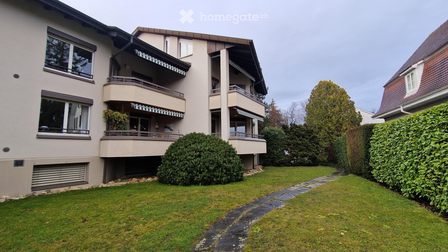 A modern two-story residential building with balconies, large windows, and a well-maintained garden with a stone pathway.